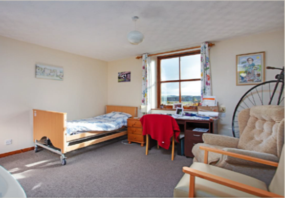 A room with a bed, desk, and chair, featuring a window with curtains and a bicycle wheel on the wall.